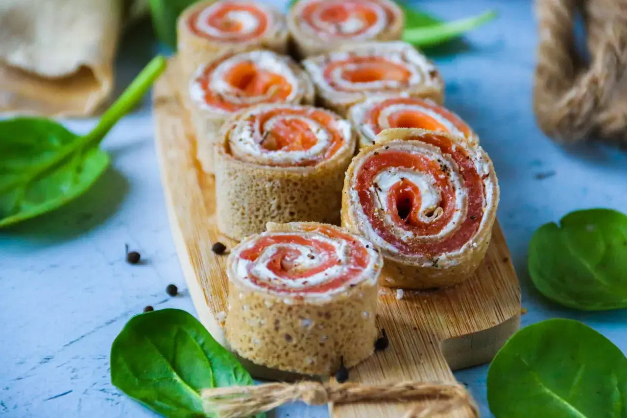 Rouleaux de cr&ecirc;pes au saumon fum&eacute; et fromage frais, parfaits pour un ap&eacute;ro gourmand. Quelques feuilles d'&eacute;pinards et grains de poivre compl&egrave;tent la sc&egrave;ne.