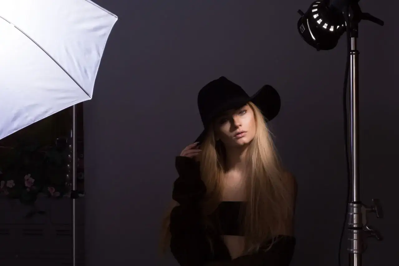 Młoda kobieta w kapeluszu, oświetlona przez lampę typu fresnel i softbox, pozuje do sesji zdjęciowej.
