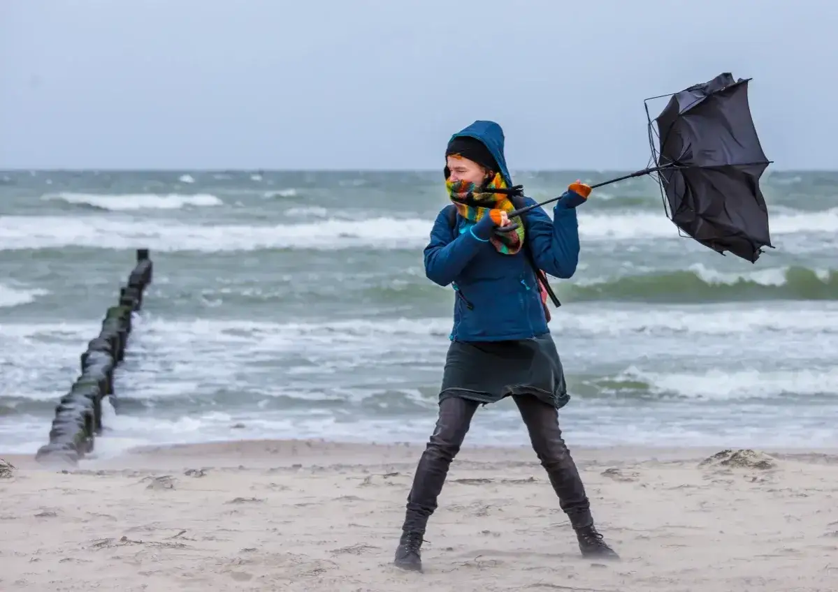 Dziewczyna walczy z wiatrem na plaży, próbując utrzymać parasolkę. Widać aktualny stan morza – wzburzone fale i pochmurne niebo.