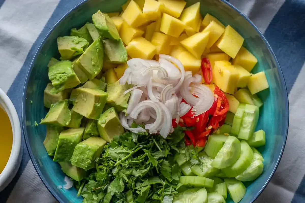 Bunte Zutaten für einen Mango-Salat: gewürfelte Mango, Avocado, Gurke, Zwiebeln, Chili und Koriander.