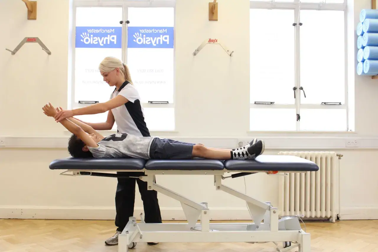 Fisioterapista assiste un bambino con ipertonia muscolare, eseguendo un esercizio di stretching per le braccia su un lettino.