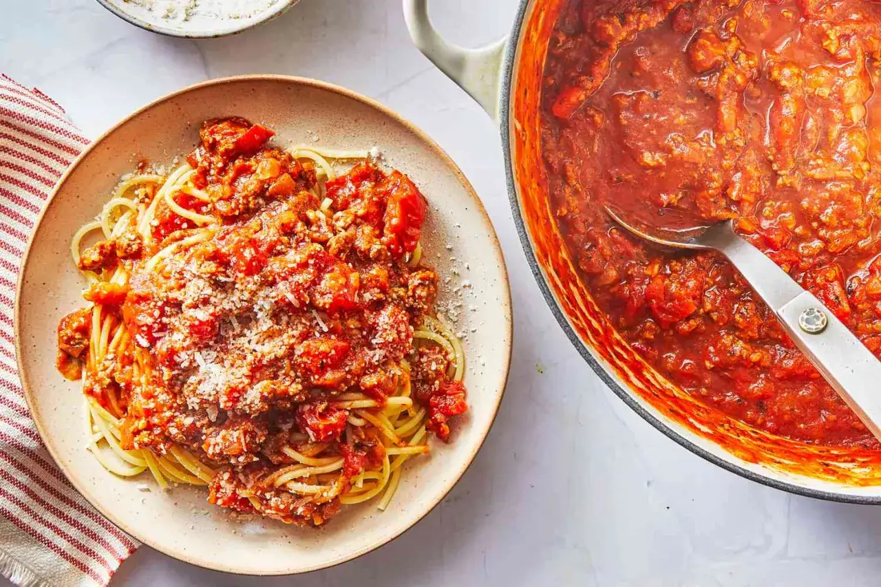 Talerz spaghetti z bogatym, mięsnym sosem do spaghetti, posypany parmezanem. Obok garnek z tym samym pysznym sosem.