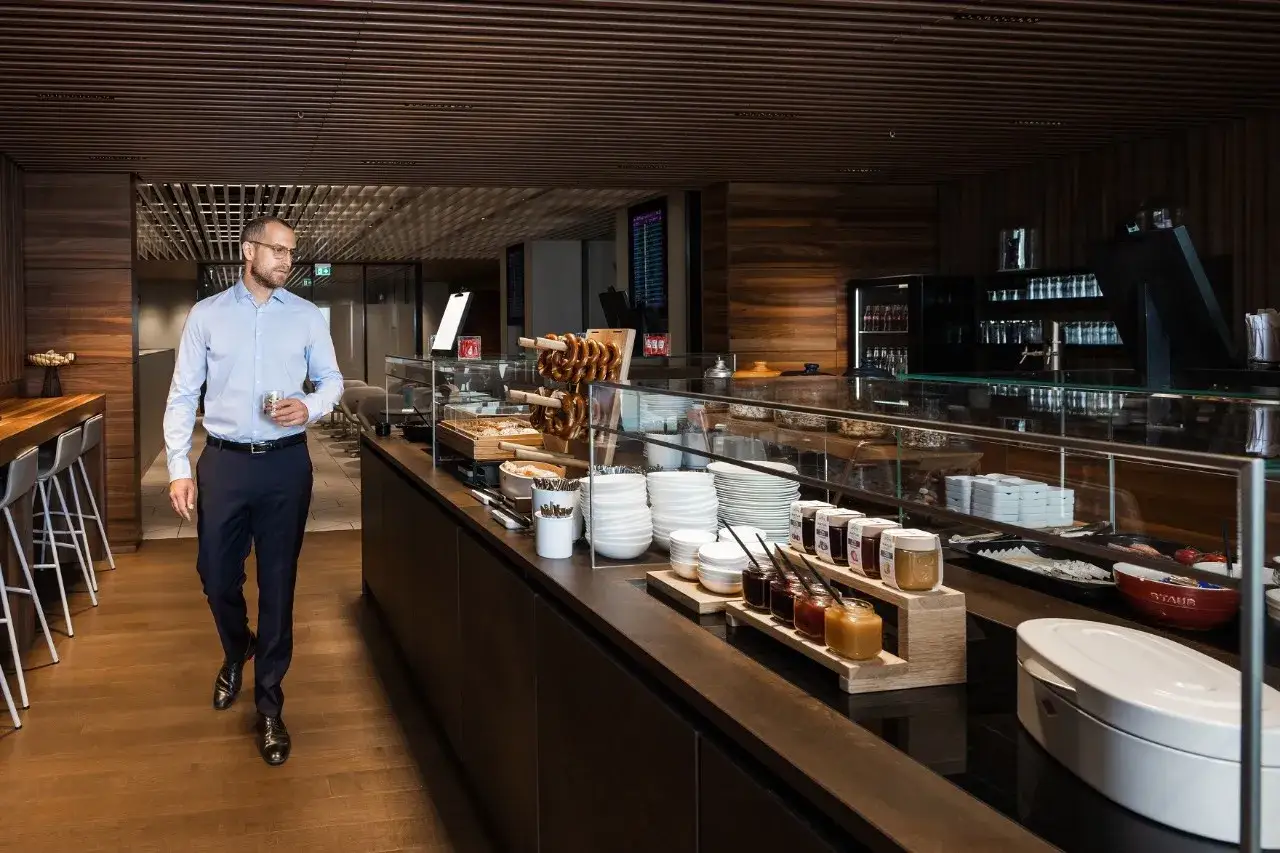 Mann in einem Anzug geht durch die **Priority Pass Lounge Berlin**, vorbei an einem Buffet mit Brezeln, Marmeladen und Geschirr.