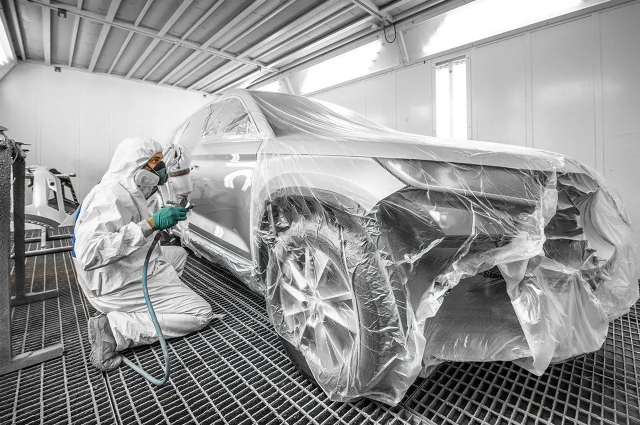 Tecnico in tuta protettiva vernicia un'auto coperta di cellophane. Consulta il listino prezzi verniciatura auto per preventivi dettagliati.