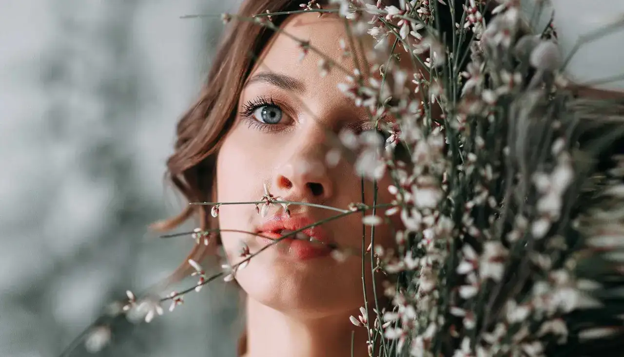 Portret fotografia młodej kobiety z niebieskimi oczami, ukrytej za gałązkami kwitnącej rośliny.