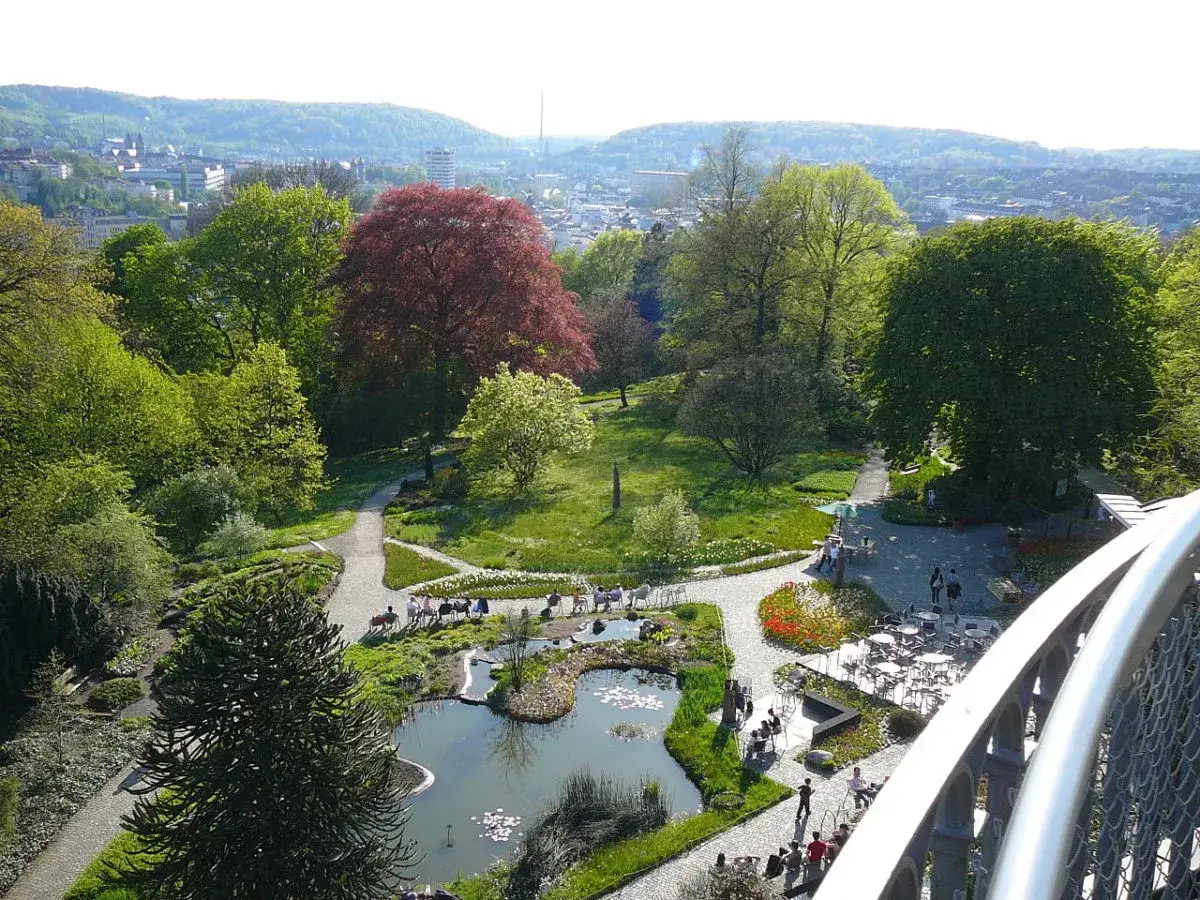 Gr&uuml;ne Oasen und bl&uuml;hende Beete in den Wuppertal Parks. Ein Teich mit Seerosen und ein roter Baum dominieren die Szenerie. Menschen genie&szlig;en die Sonne.