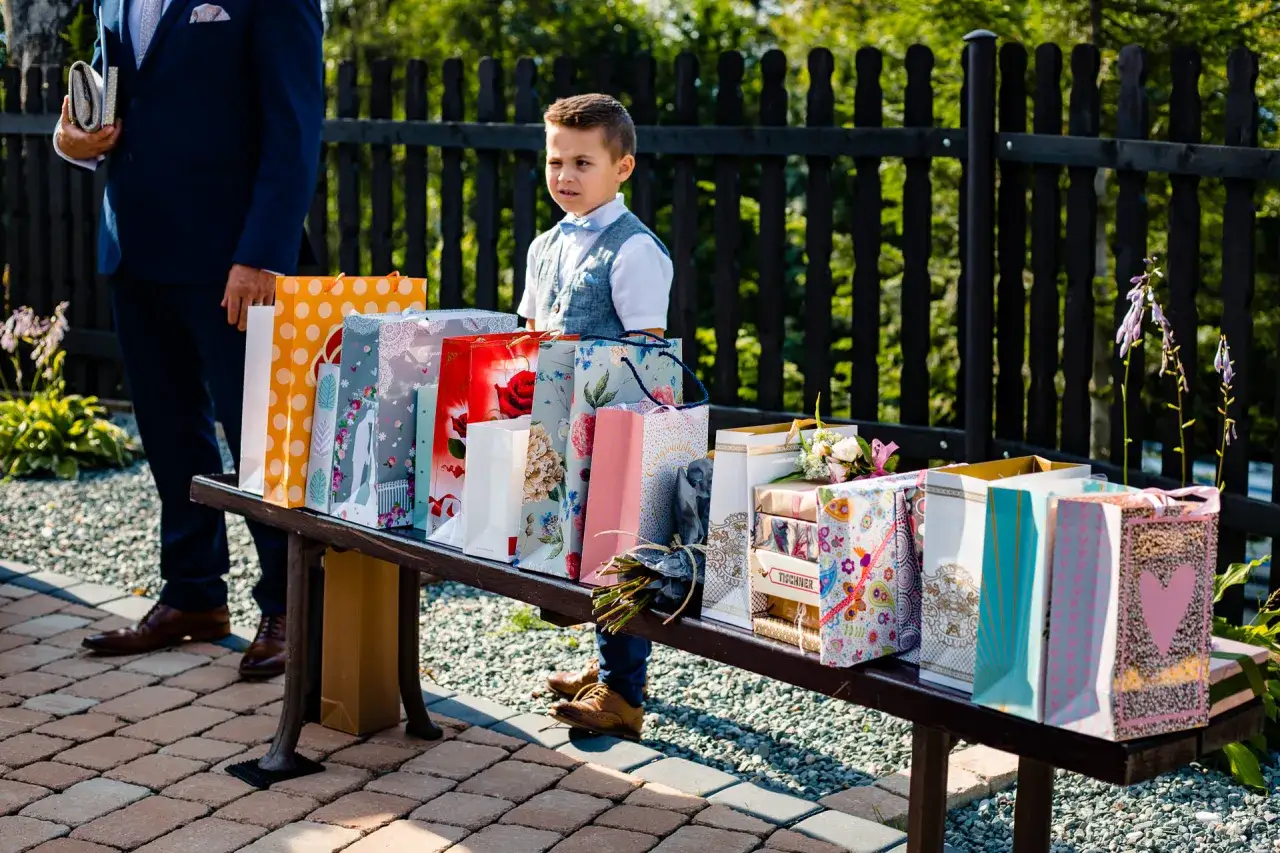 Chłopiec w eleganckim stroju stoi obok stołu pełnego prezentów, pokazując, co zamiast kwiatów na ślub może być świetnym pomysłem.