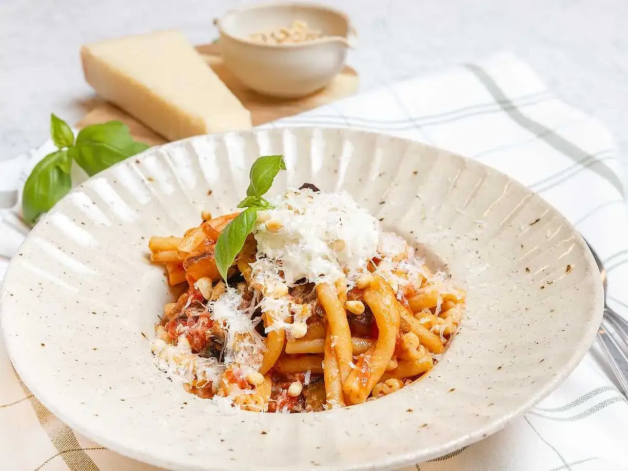 En portion pasta alla norma med aubergine, tomatsås och toppad med ricotta och pinjenötter. En bit parmesan och färsk basilika i bakgrunden.