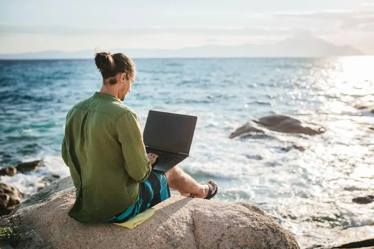 Hombre con port&aacute;til en roca junto al mar, disfrutando de trabajos para n&oacute;madas digitales.