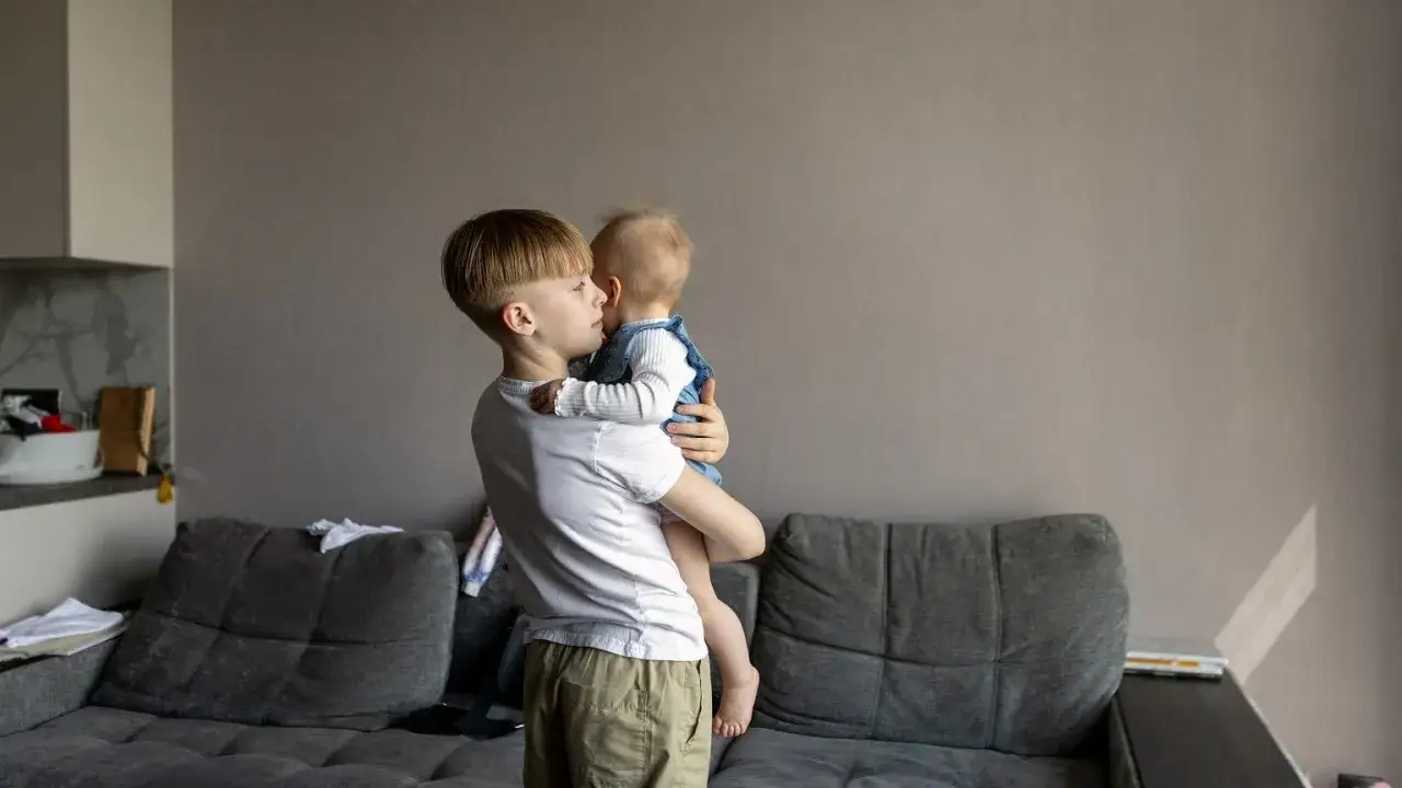 Starszy brat trzyma na rękach niemowlę. Zastanawiasz się, od jakiego wieku dziecko może zostać samo w domu?