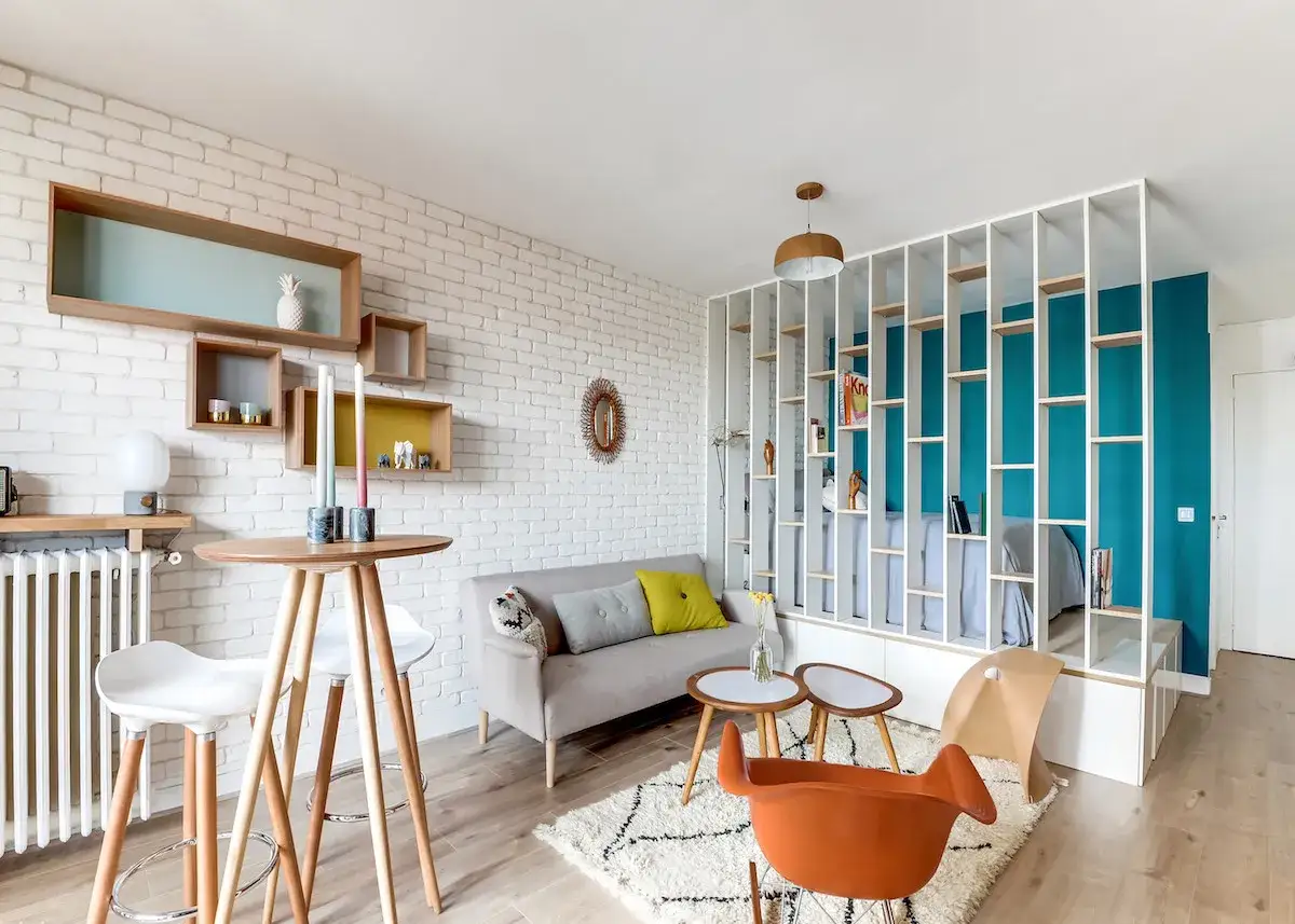 Aménagement studio 25 m2 : salon cosy avec canapé gris, table haute, étagères murales et un lit séparé par une bibliothèque ajourée.