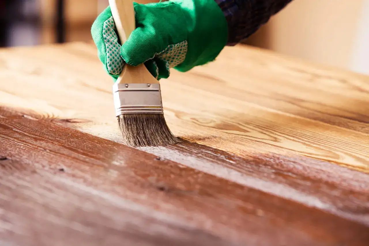 Main d'une personne portant un gant vert, appliquant du vernis sur un meuble en bois avec un pinceau.