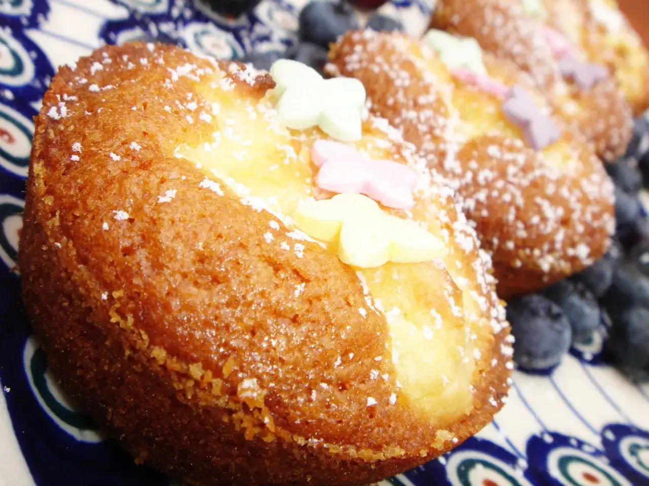 Puszyste muffinki serowe, posypane cukrem pudrem i ozdobione kolorowymi motylkami, leżą obok świeżych borówek na talerzu.