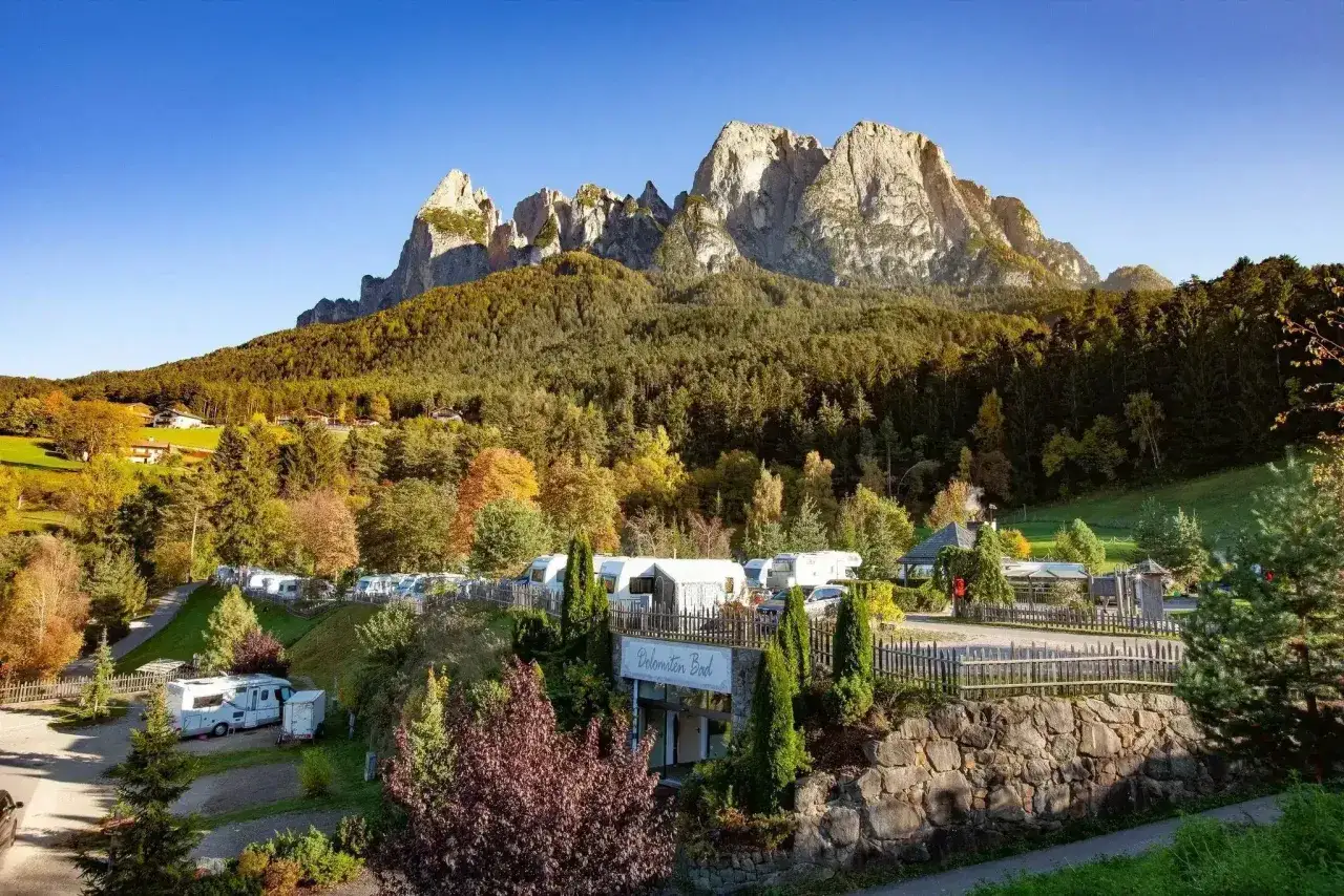 Malowniczy camping Seiser Alm z kamperami i przyczepami, otoczony jesiennym lasem i majestatycznymi Dolomitami.
