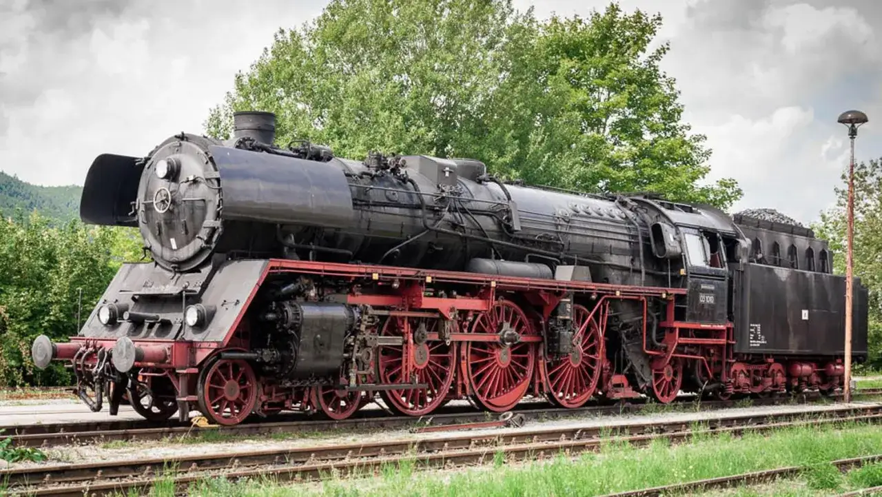 Eine historische Dampflokomotive, ein Symbol der Erfindung Eisenbahn, steht auf Gleisen vor gr&uuml;nen B&auml;umen.