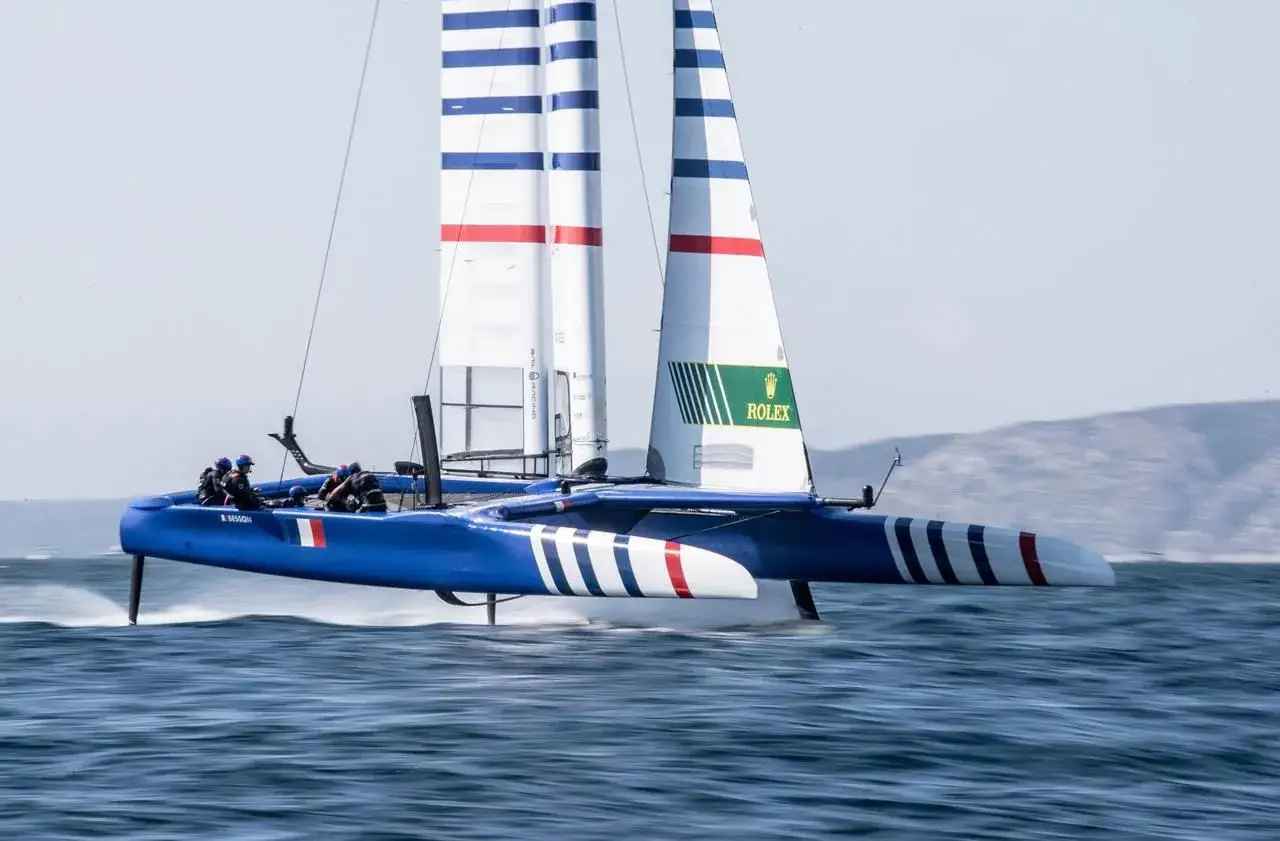 Catamaran bleu et blanc, voiles rayées, en pleine action. La **vitesse catamaran** est impressionnante, les foils hors de l'eau.