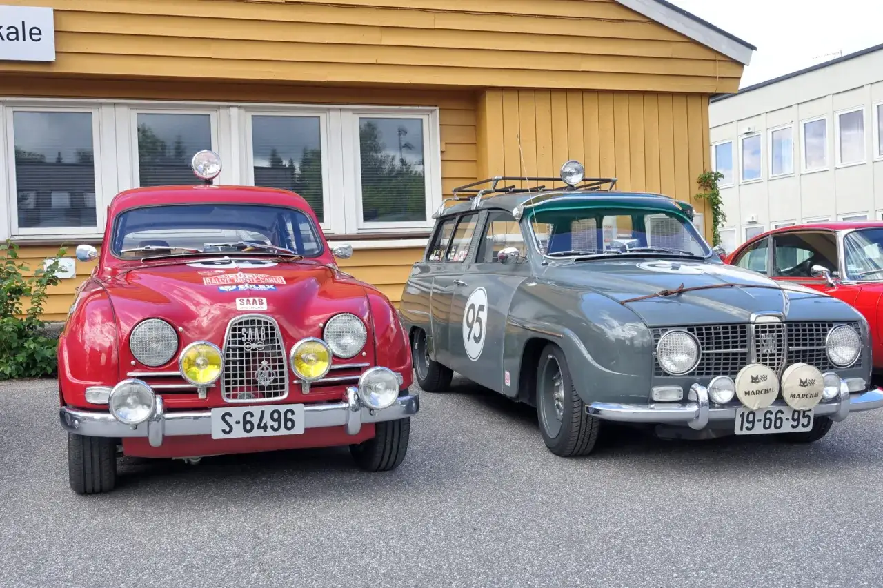 Tv&aring; klassiska Saab-bilar, en r&ouml;d och en gr&aring;, st&aring;r parkerade framf&ouml;r en gul byggnad. Den r&ouml;da bilen har texten 