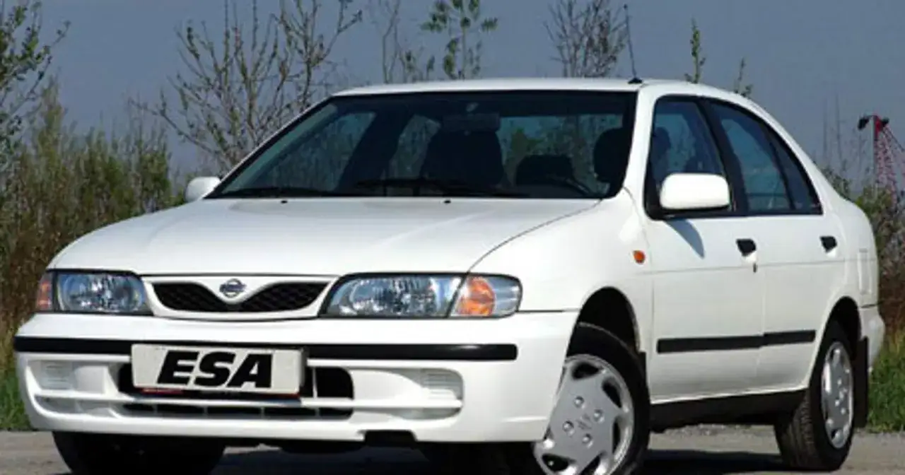 Biały Nissan Almera z lat 90. stoi na tle krzewów. Z przodu widoczna tablica rejestracyjna z napisem