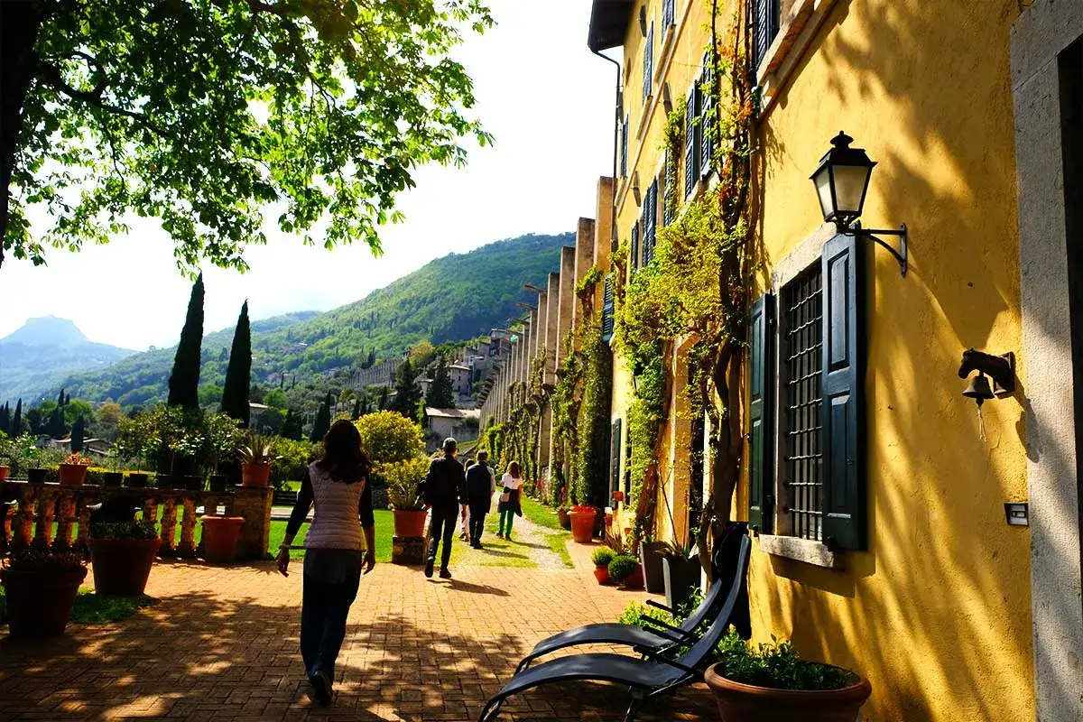 Passeggiata in una soleggiata limonaia la malora, con persone che camminano lungo un muro giallo ricoperto di rampicanti e un paesaggio collinare sullo sfondo.
