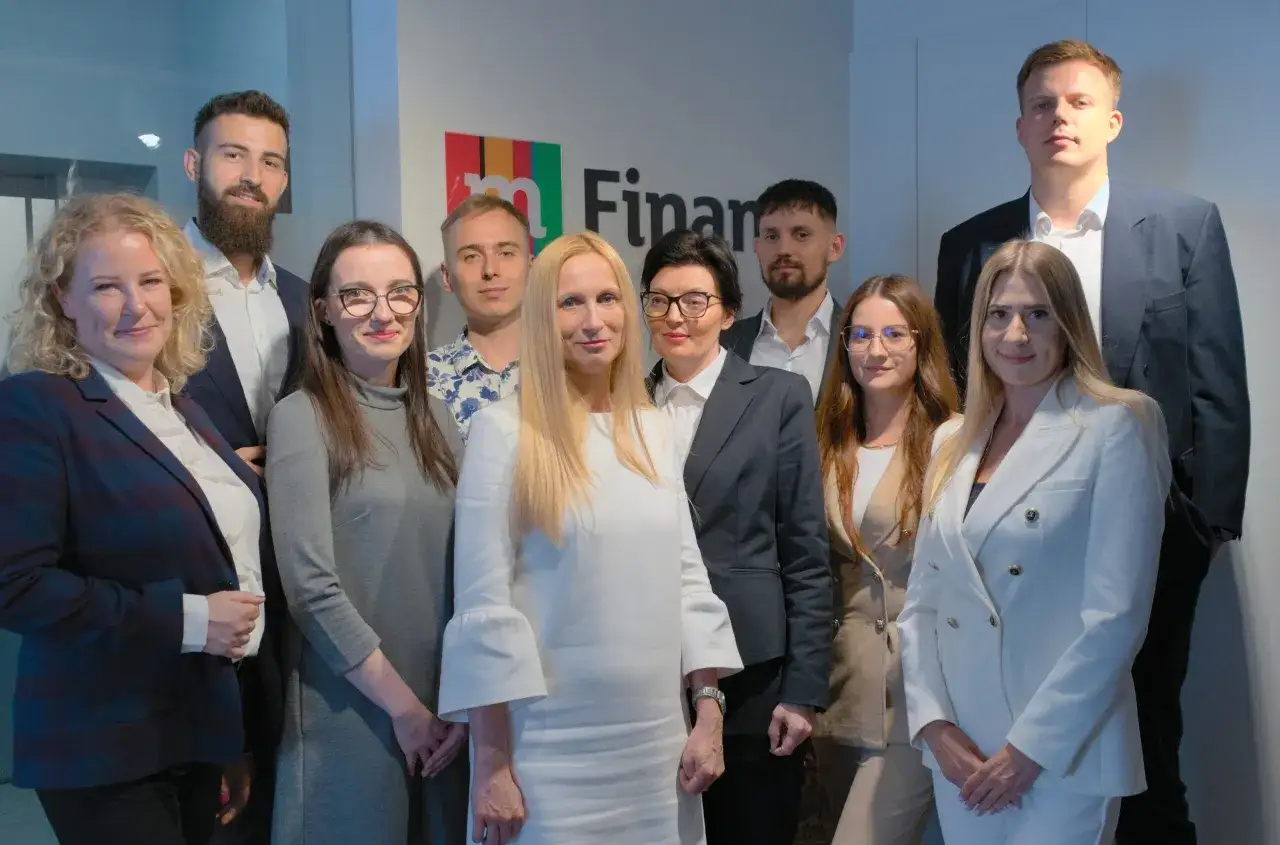 Grupa profesjonalistów w garniturach i strojach biznesowych pozuje przed logo firmy.