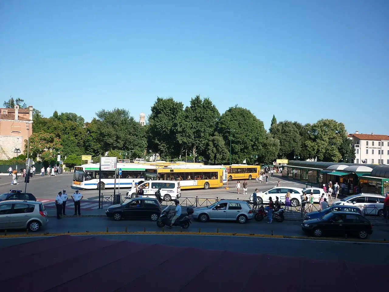 Traffico e persone a **Piazza Roma, Venezia**. Autobus gialli e bianchi, auto, moto e agenti in divisa animano la scena sotto un cielo azzurro.