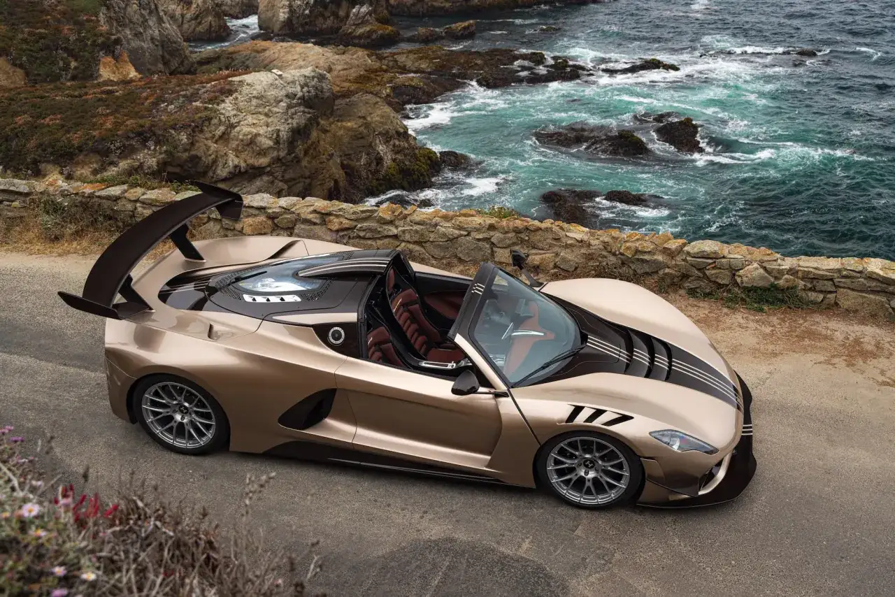 Ein Hennessey Venom F5 Roadster in Goldbraun steht an einer felsigen K&uuml;ste. Das offene Verdeck gibt den Blick auf braune Ledersitze frei.