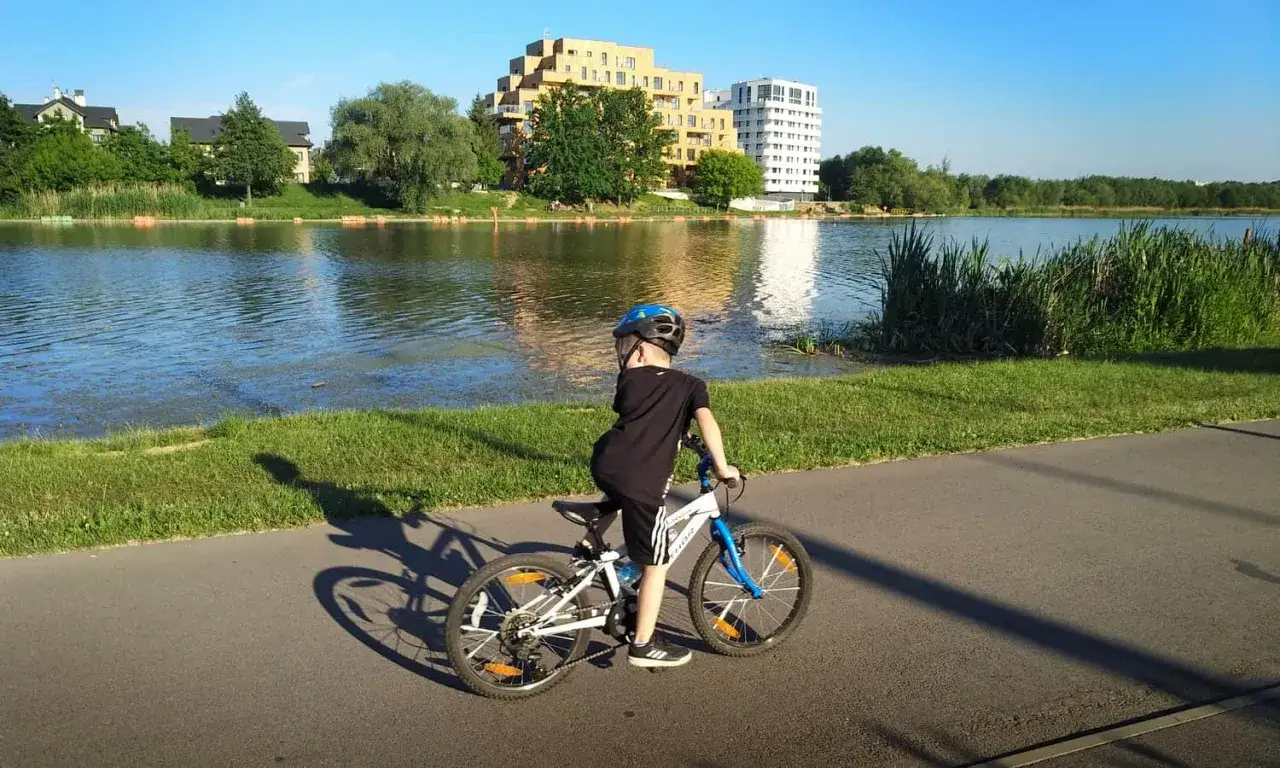 Chłopiec na rowerze cieszy się pięknymi trasami rowerowymi Rzeszów nad jeziorem, z nowoczesnymi budynkami w tle.