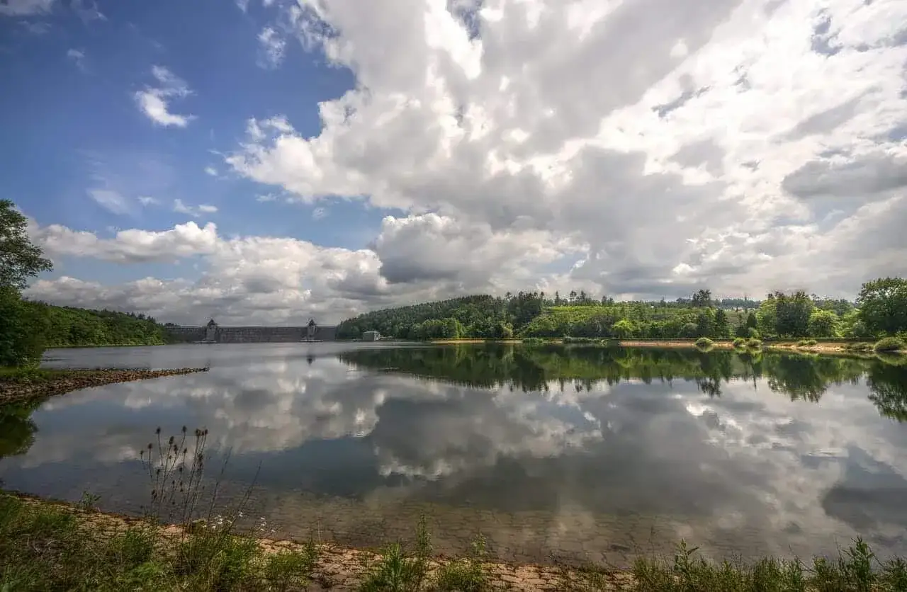 Stausee mit imposanter Staumauer, gespiegelt im ruhigen Wasser. Grüne Hügel und eine dramatische Wolkenformation sind ebenfalls zu sehen, ein malerischer Anblick, wie er in NRW oft zu finden ist.