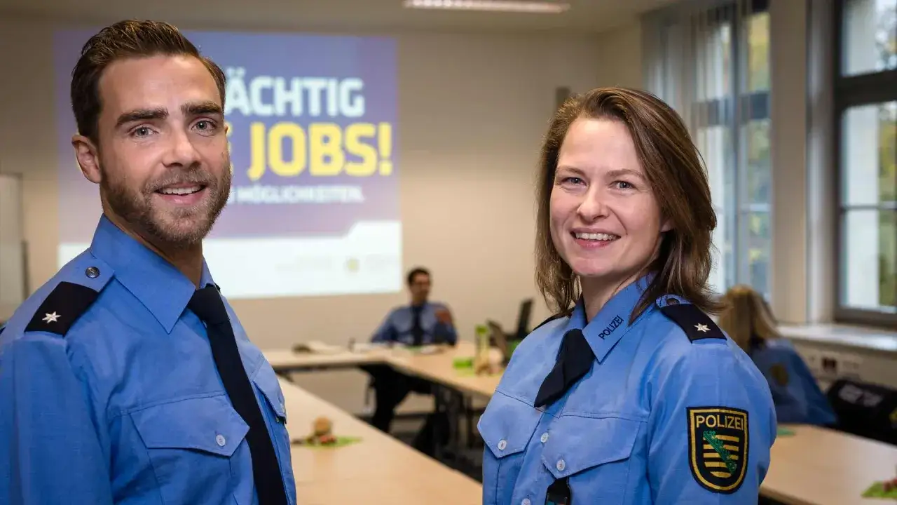 Zwei Polizisten l&auml;cheln in die Kamera. Im Hintergrund eine Leinwand mit 
