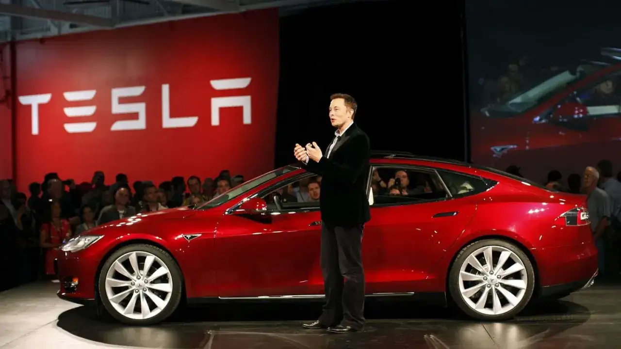 Elon Musk présente une Tesla rouge vif sur scène, devant le logo de la marque et un public captivé.