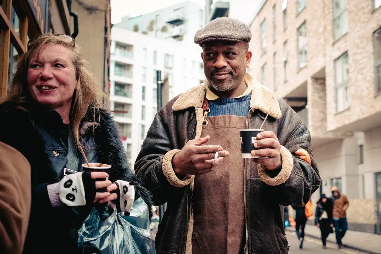 A man in a flat cap and shearling jacket holds a coffee, a perfect subject for a best Fuji lens for street photography.