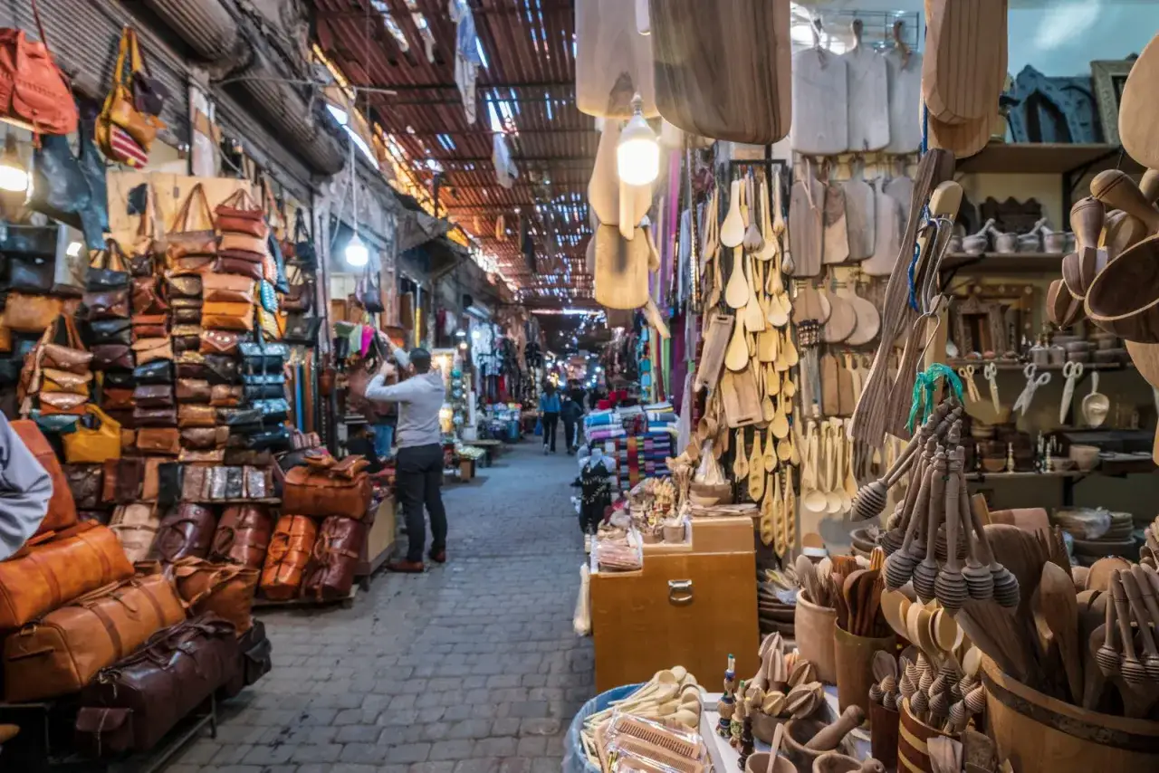 Un homme examine des sacs en cuir dans un souk anim&eacute; de Marrakech, entour&eacute; d'&eacute;tals d&eacute;bordant d'ustensiles en bois et d'artisanat.
