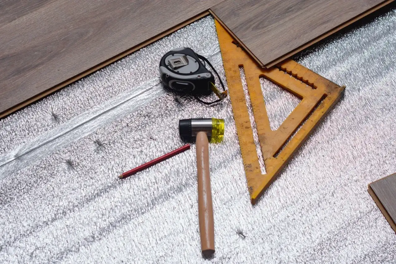 Podkład pod panele laminowane: taśma miernicza, młotek, ołówek i ekierka leżą na srebrnej piance izolacyjnej.