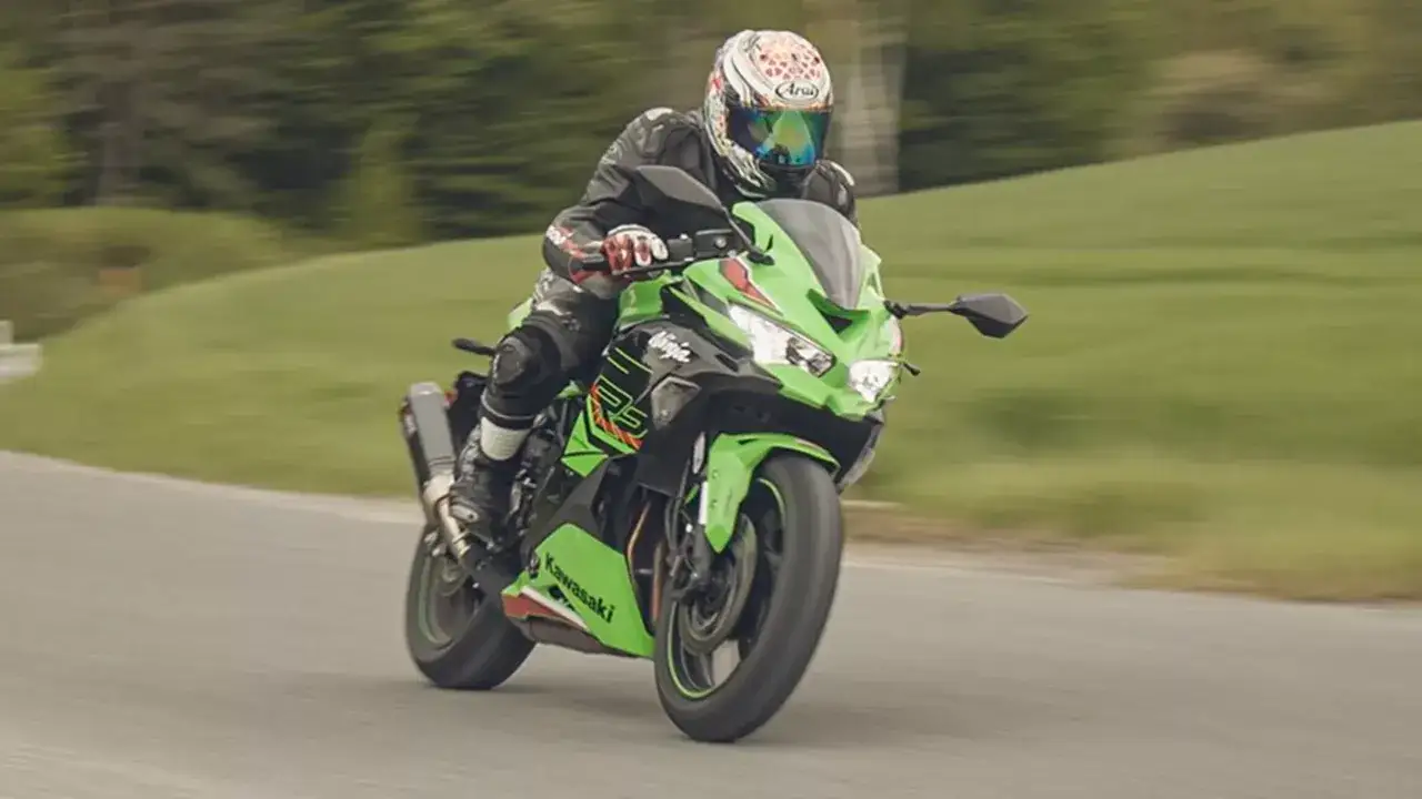 Motocyklista w kasku i kombinezonie pędzi na zielonym motocyklu Kawasaki zx4rr po krętej drodze wśród zielonych wzgórz.
