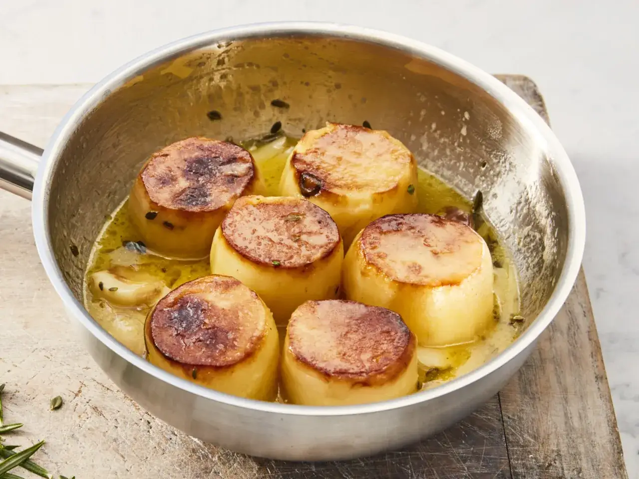 Sechs goldbraune Fondantkartoffeln brutzeln in einer Pfanne in aromatischer Butter mit Kr&auml;utern und Knoblauch.