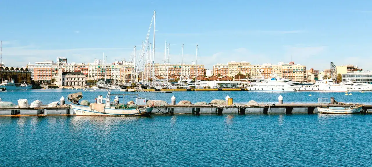 Barcos de pesca y yates de lujo atracados en la marina valencia, con edificios coloridos de fondo bajo un cielo azul.