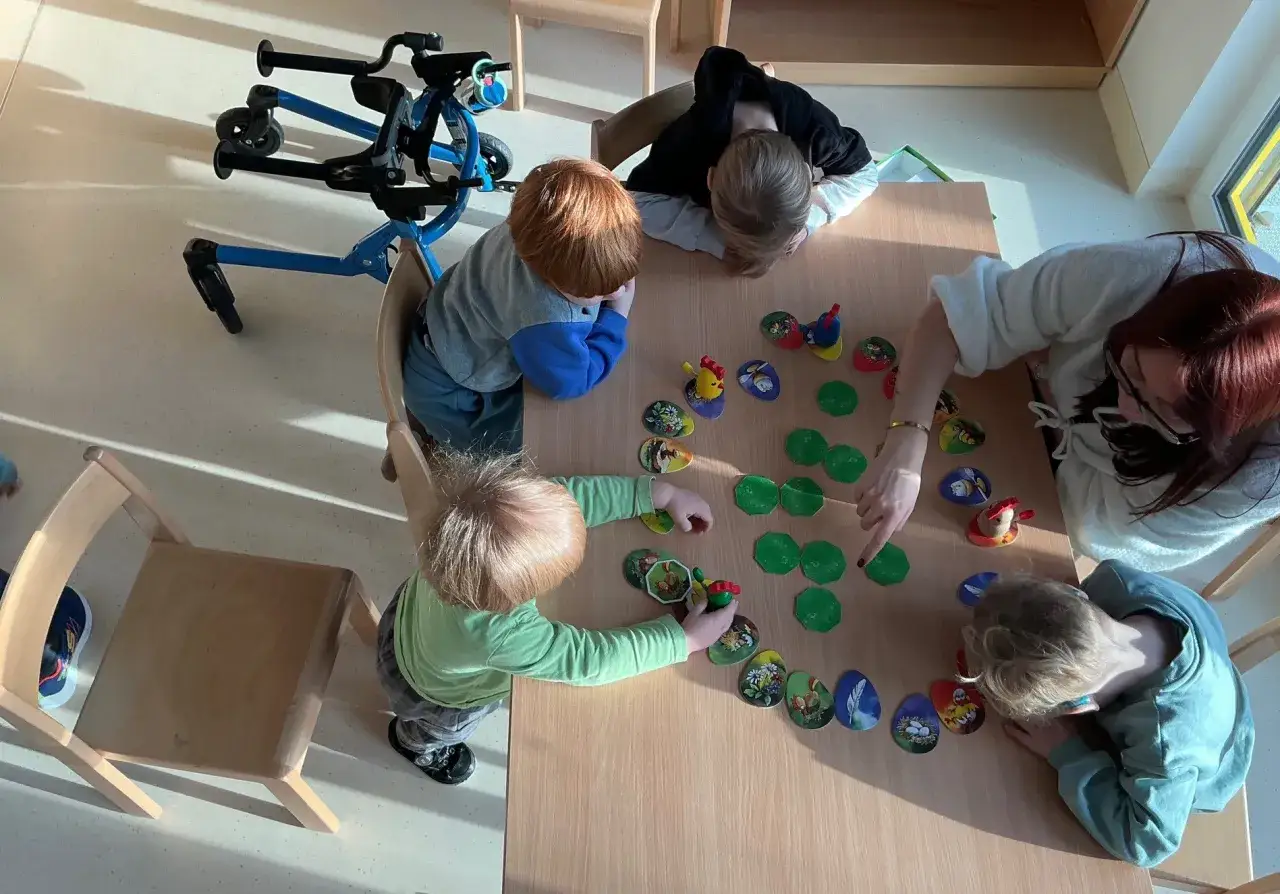 Kinder spielen und lernen gemeinsam am Tisch. Ein Gehwagen steht bereit. Inklusion im Kindergarten wird hier gelebt.