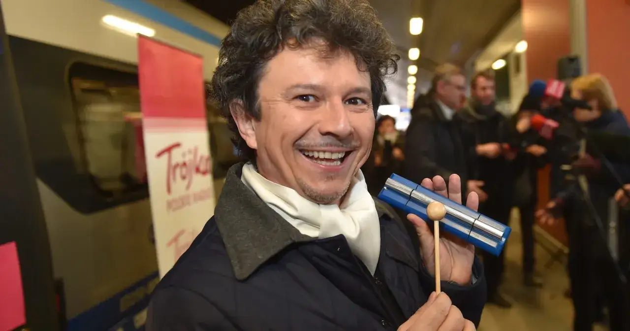 Uśmiechnięty Jacek Wójcicki, znany ze swojego charakterystycznego wzrostu, prezentuje instrument muzyczny na tle pociągu i mediów.