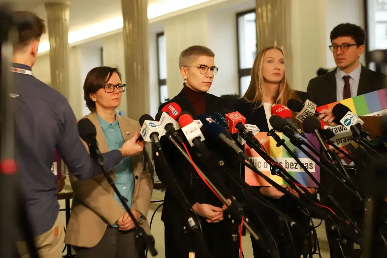 Trzy osoby udzielają wywiadu mediom, otoczone przez mikrofony. W tle tęczowa tęcza i hasło