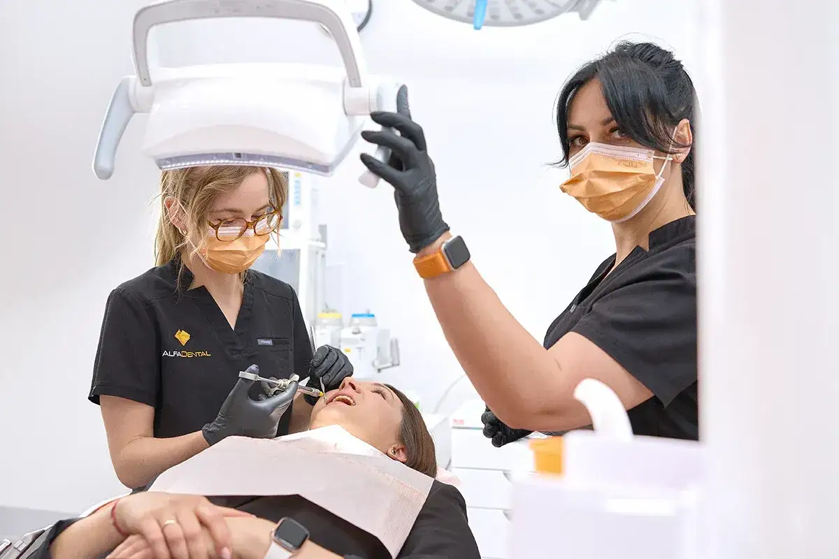 Dentystka w masce wykonuje borowanie zęba pacjentce. Druga dentystka asystuje, regulując lampę.