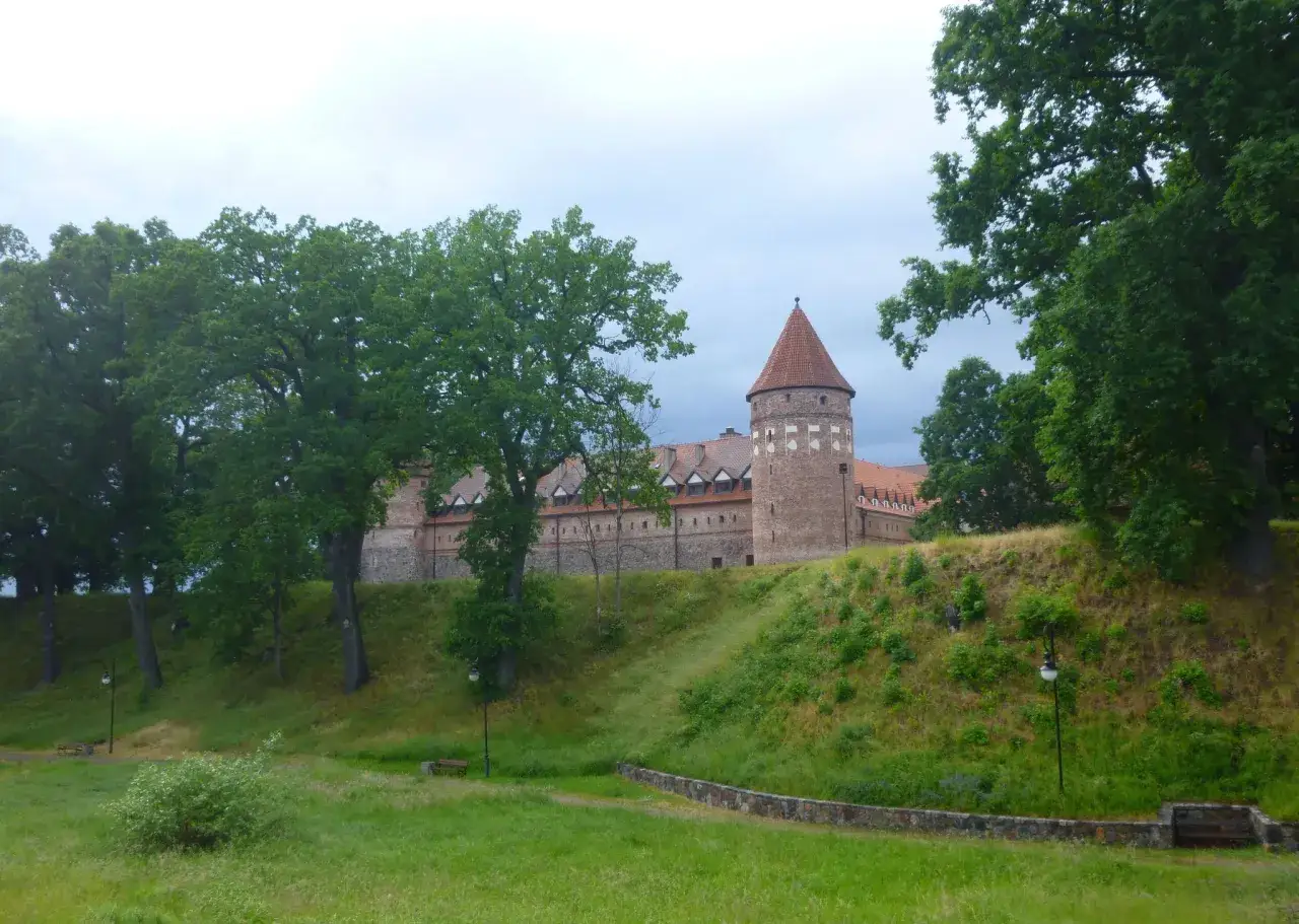 Zamek z cegły z charakterystyczną wieżą, otoczony zielenią. Idealne miejsce na spacer, tworzące malowniczą bytów trasę.