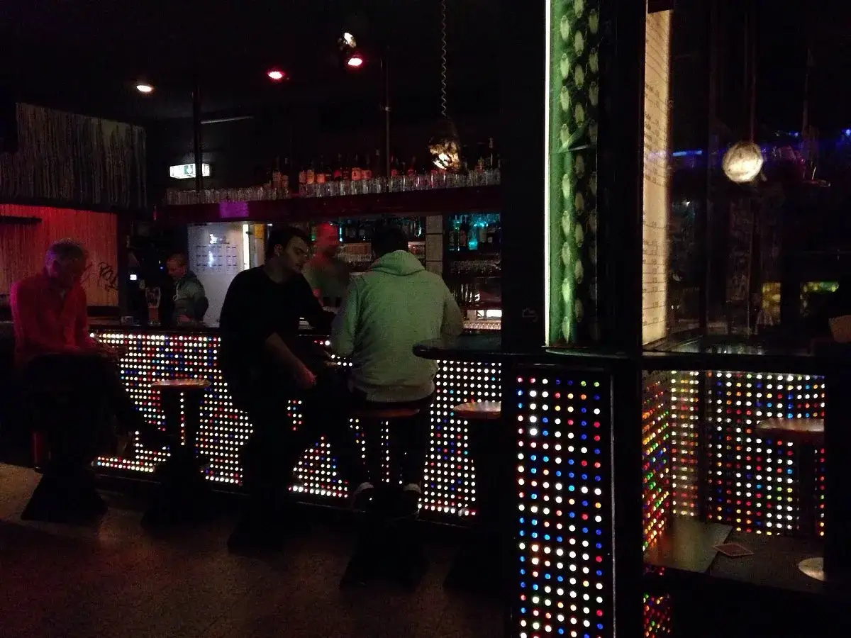 Leute sitzen an einer Bar mit bunten Lichtern in Stuttgart. Ein Mann in Rot, ein Mann in Schwarz und ein Mann in Gr&uuml;n.