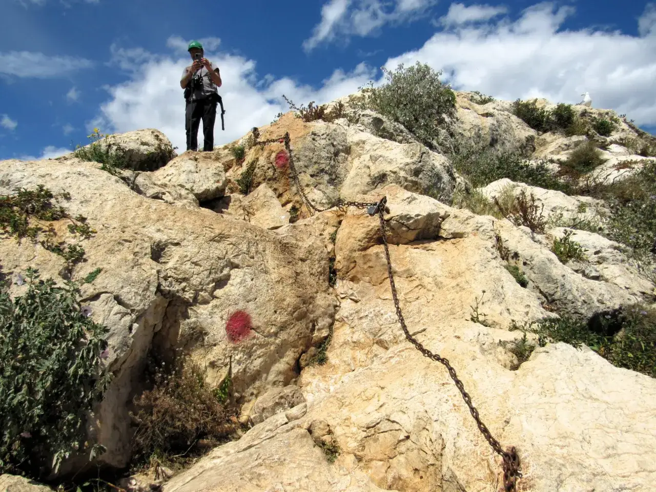 Persona con gorra verde y mochila se prepara para subir al Peñón de Ifach, usando cadenas para el ascenso.
