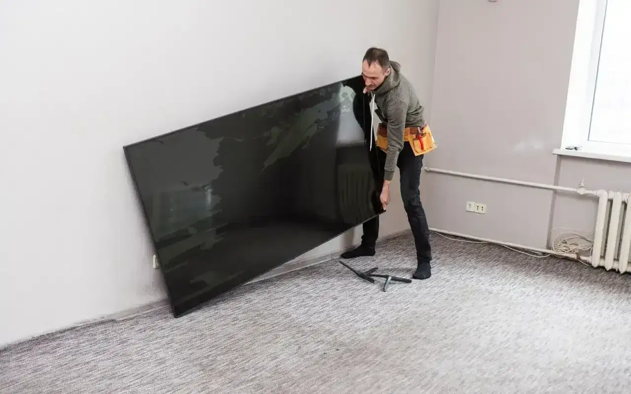 Mężczyzna z pasem narzędziowym przy ścianie trzyma duży telewizor, zastanawiając się, na jakiej wysokości powiesić TV, by był idealnie widoczny.