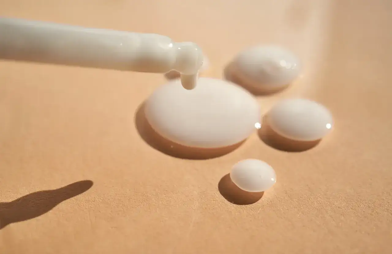 Tropfen einer weißen Flüssigkeit, die aus einer Pipette auf eine beige Oberfläche fallen. Das ist Panthenol, ein Wirkstoff für Haut und Haar.