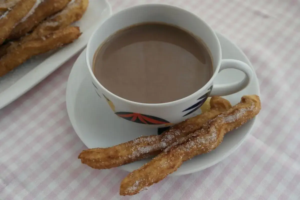 Churros caseros reci&eacute;n hechos, espolvoreados con az&uacute;car, junto a una taza de chocolate caliente.