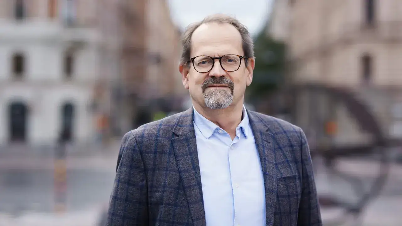 Peder Hasslev, en man med glas&ouml;gon och sk&auml;gg, b&auml;r en rutig kavaj och ljusbl&aring; skjorta. Bakgrunden &auml;r en suddig stadsmilj&ouml;.