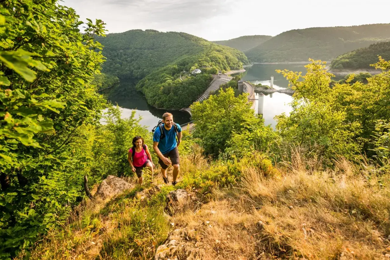 Couple randonnant sur un sentier verdoyant, avec un barrage et un lac en arrière-plan, dans le parc national de l'Eifel.