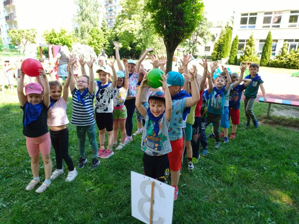 Radosne dzieci biorą udział w konkurencjach sportowych dla dzieci, trzymając balony i machając rękami.