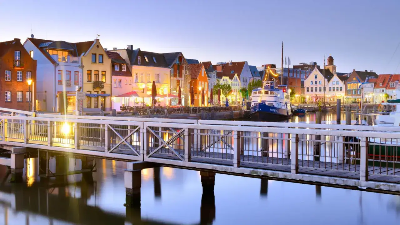 En bro leder till en charmig hamn i Husum, Tyskland, med f&auml;rgglada hus och b&aring;tar som speglas i vattnet.
